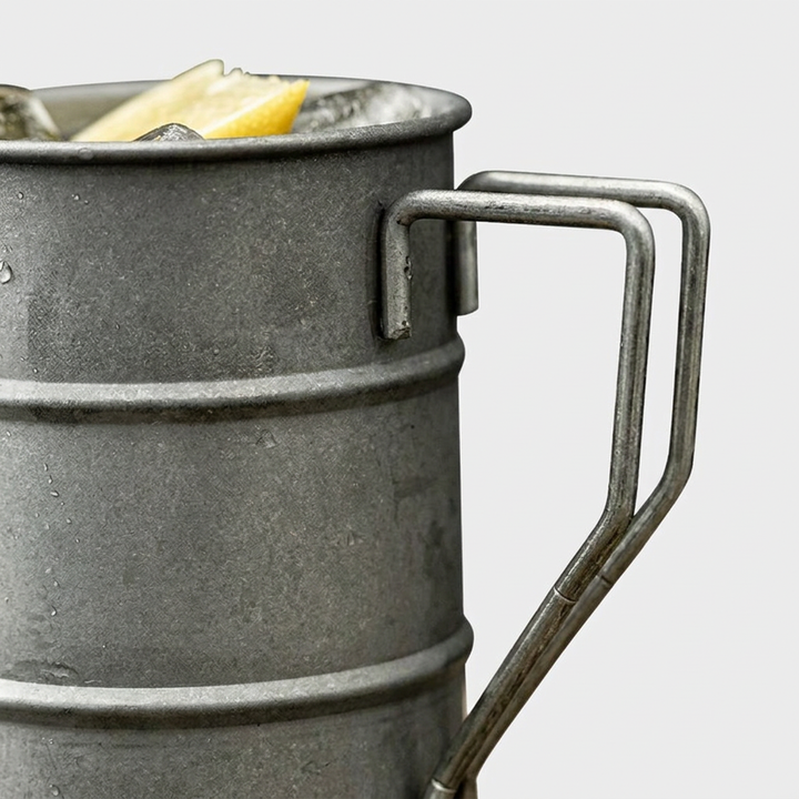 Close-up of a galvanized metal bucket-style drink holder with a double-pronged handle, shown filled with ice and a lemon slice against a light gray background.