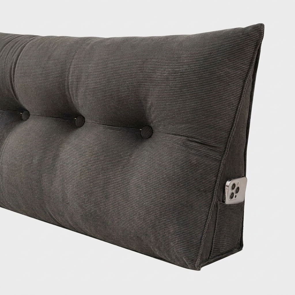 A close-up of a dark gray corduroy headboard with button tufting and a side pocket holding a silver smartphone, set against a plain light gray background.